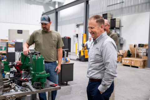 Two men in a workshop stand near a table with green machinery parts. One man in a gray shirt smiles while the other, sporting a cap and green t-shirt, demonstrates equipment—reflecting hands-on Lean Manufacturing Consulting amid tools and machinery.