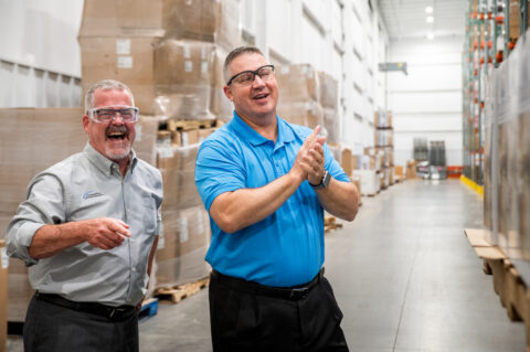 Two men in safety glasses and business casual attire stand in a warehouse, smiling and laughing. One claps while the other gestures, embodying operational excellence amid stacks of large boxes and pallets in the background.