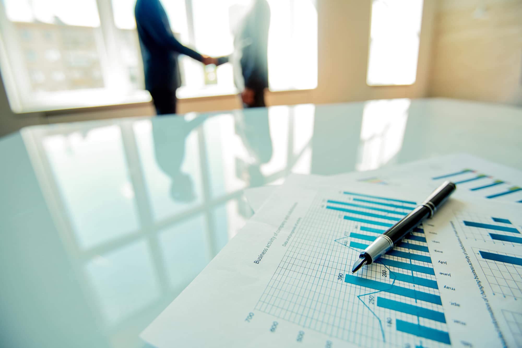 A close-up of financial charts and a pen on a glass table, with two blurred people shaking hands in the background in a bright office setting.