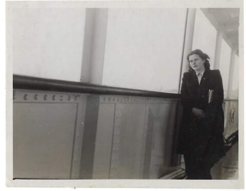 A woman in a dark coat stands alone, leaning against a metal railing on a ship’s deck, holding a book or folder, gazing thoughtfully into the distance. The photo is black and white.