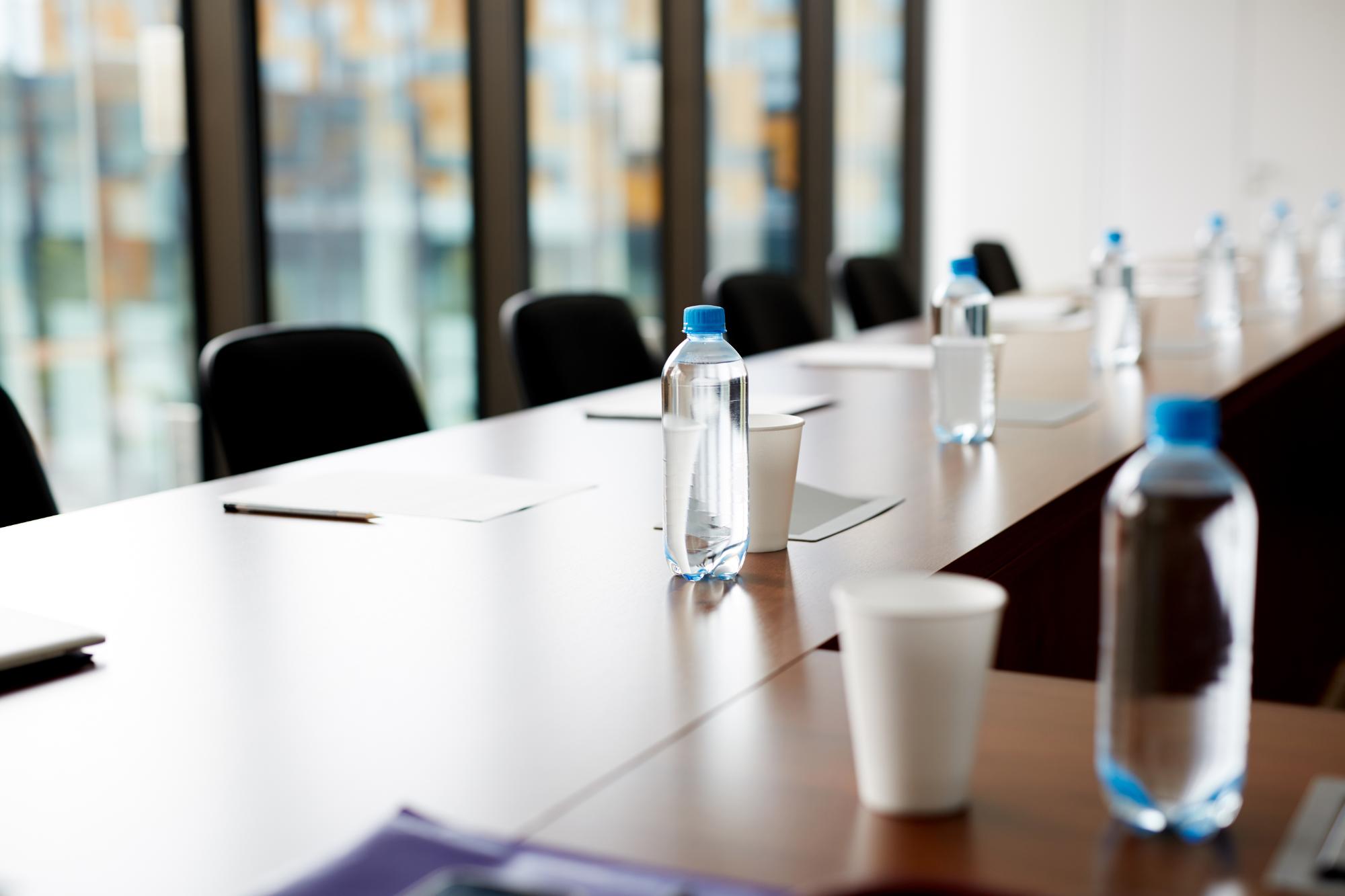 A long conference table with black chairs, each place set for a meeting focused on operational excellence, featuring water bottles, paper cups, pens, and sheets of paper. Large windows in the background let in natural light.