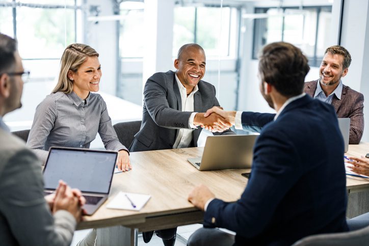 Four business professionals sit around a conference table with laptops, while two of them reach across the table to shake hands and smile, highlighting a successful aerospace and defense consulting meeting or agreement.