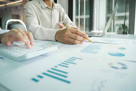Two people work at a desk with charts, graphs, and a laptop. One person uses a calculator while the other points at a document with a pencil, suggesting financial or data analysis.